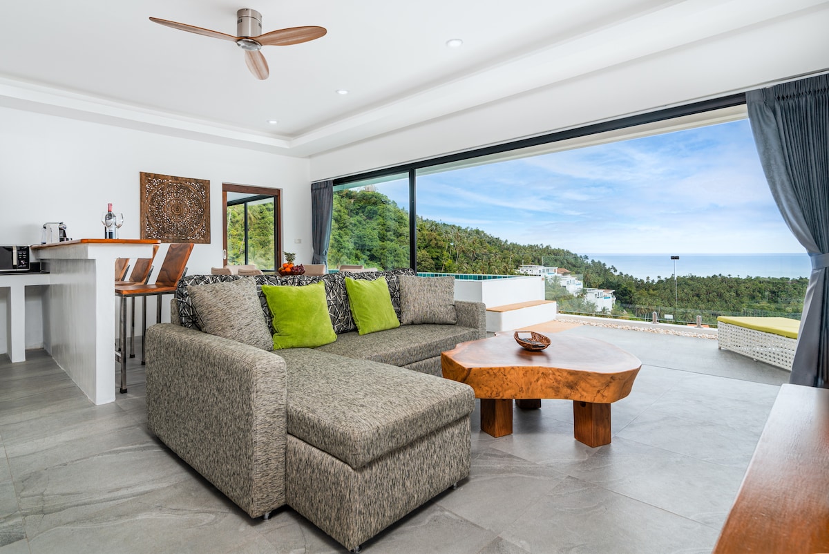 A spacious living area is presented, featuring a large sectional sofa adorned with green accent pillows. A natural wood coffee table is positioned in front of the sofa. Floor-to-ceiling sliding glass doors provide an unobstructed view of the lush hillside and expansive sky.