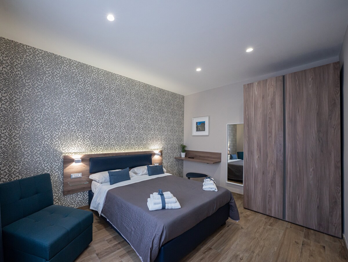 A modern room features a large bed with a neutral-colored blanket and decorative towels arranged at the foot. A cozy armchair sits beside the bed, while a wardrobe and a mirror are placed against the wall. Soft lighting enhances the elegant patterned wallpaper.