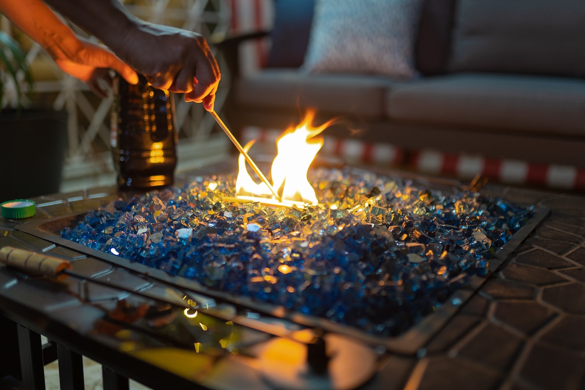 A fire pit is surrounded by sparkling blue glass stones, with flames flickering above. The scene is complemented by a cozy seating area in the background, featuring a comfortable sofa and decorative pillows.