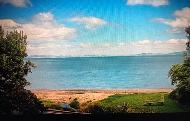 A serene coastal view is presented, featuring a sandy beach leading to calm blue waters. Lush green vegetation borders the scene, with gentle waves visible in the distance under a partly cloudy sky.