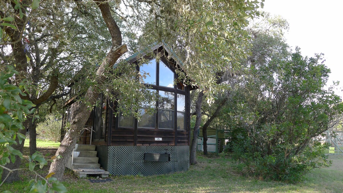 The tiny house features large front windows, allowing natural light to illuminate the interior. Surrounded by trees, the exterior showcases wood paneling and a staircase leading to the entrance. A peaceful outdoor setting is enhanced by greenery and open space.