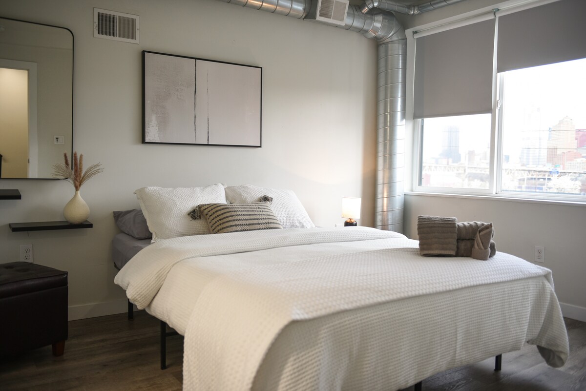 חדר שינה מעוצב עם אסתטיקה מודרנית, עם תעלות חשופות, חלונות גדולים ומצעים רכים ושכבות.