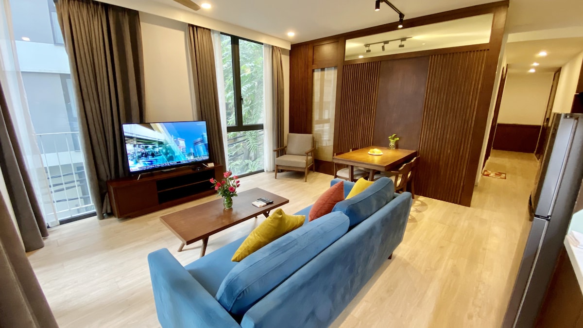 A spacious living area is highlighted by a blue sofa with colorful cushions, a wooden coffee table, and a flat-screen TV. Large windows allow natural light to fill the room, while elegant curtains frame the view. A dining table and nearby hallway lead towards other areas of the apartment.