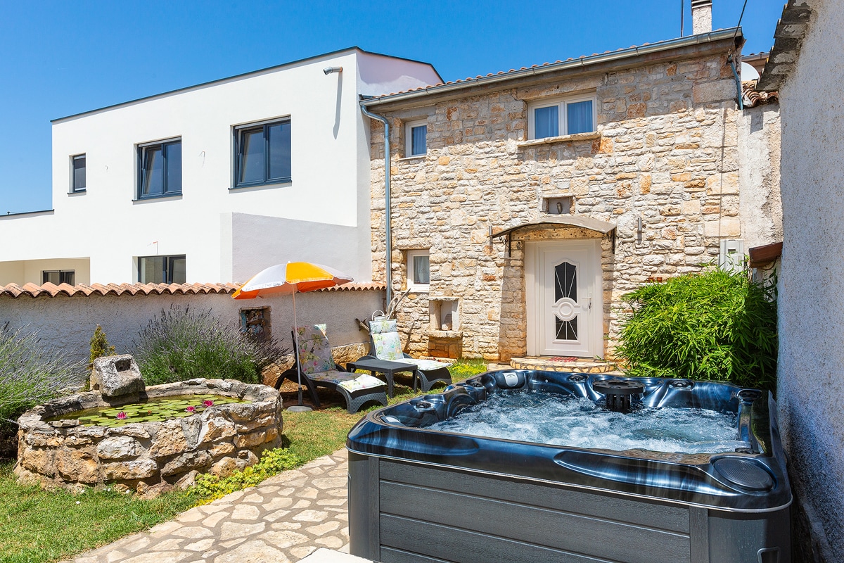 A fenced outdoor courtyard features a black hot tub, surrounded by lush greenery and stone pathways. A stone wall and a small garden area add to the charm. Nearby, a sunbathing area is equipped with loungers and an umbrella, creating a relaxing outdoor retreat.