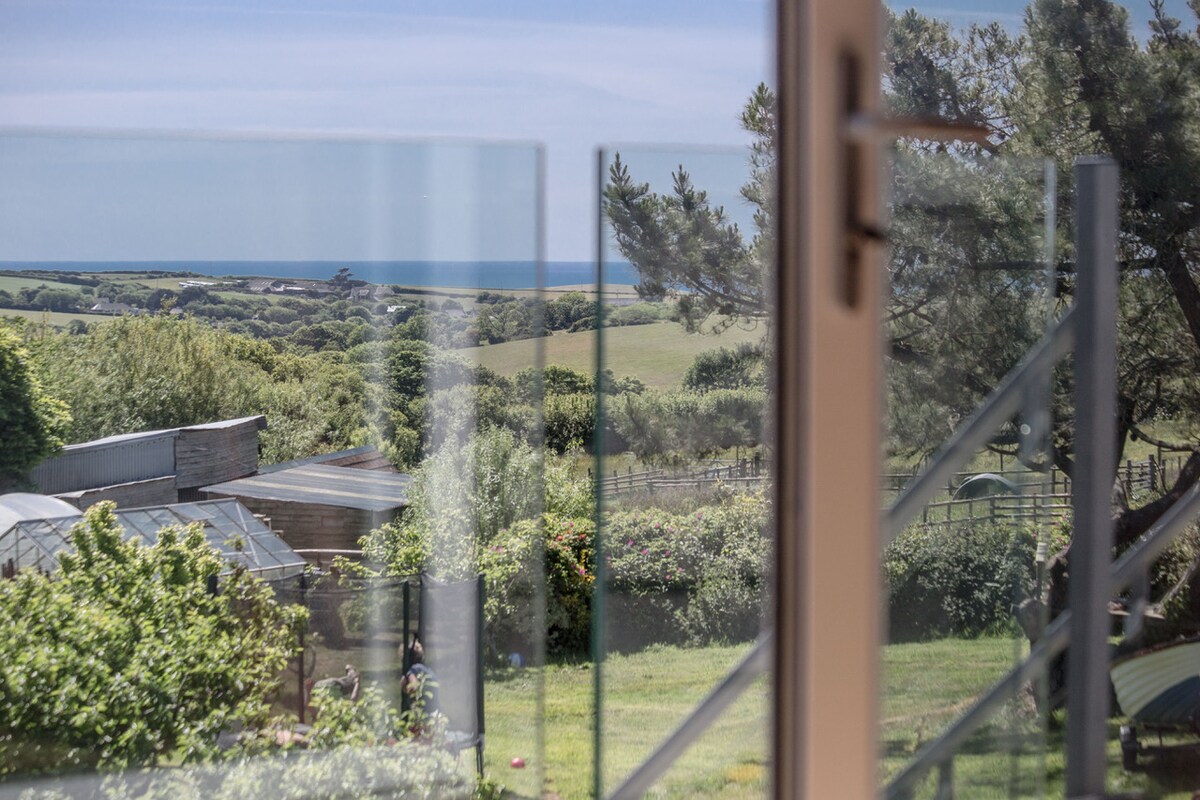 A view of lush greenery and rolling hills extends towards the coastline, visible through a glass panel. The scene captures a serene landscape with a blue sky above, framed by trees in the foreground.