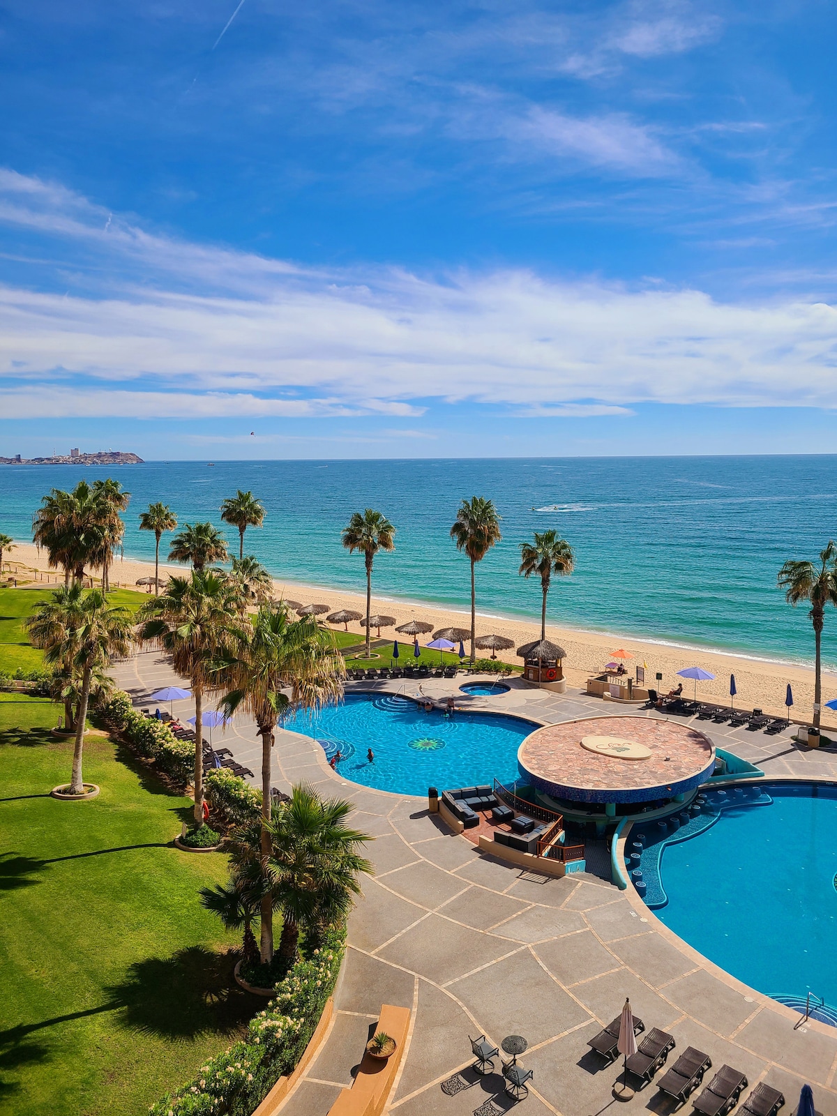 A sweeping view of a beachfront area is visible, featuring lush green lawns and palm trees lining the shore. A large swimming pool with a circular island bar is positioned nearby, connecting effortlessly to the tranquil ocean beyond.