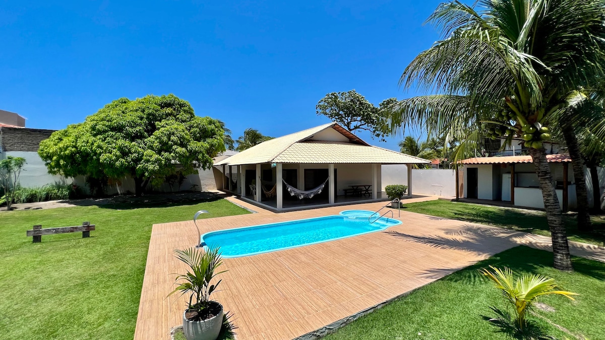 The outdoor area features a fenced swimming pool surrounded by a wooden deck. A shaded patio offers seating areas, complemented by palm trees and lush green grass. Hammocks are suspended near the house, providing a relaxed atmosphere for leisure.