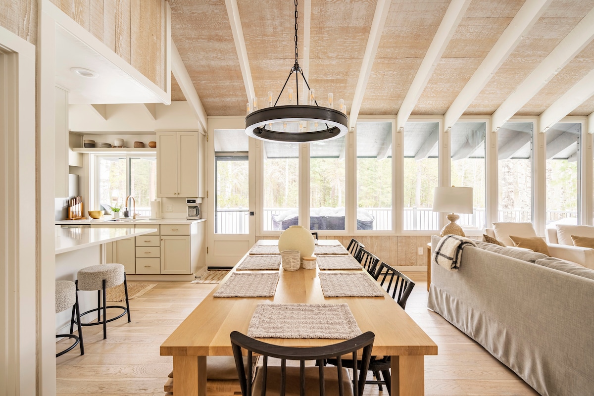 An open-concept dining area features a large wooden table with black chairs, set for a meal. Light fills the space through expansive windows, showcasing nature outside. A stylish chandelier hangs from a beamed ceiling, and a comfortable living area is visible in the background.