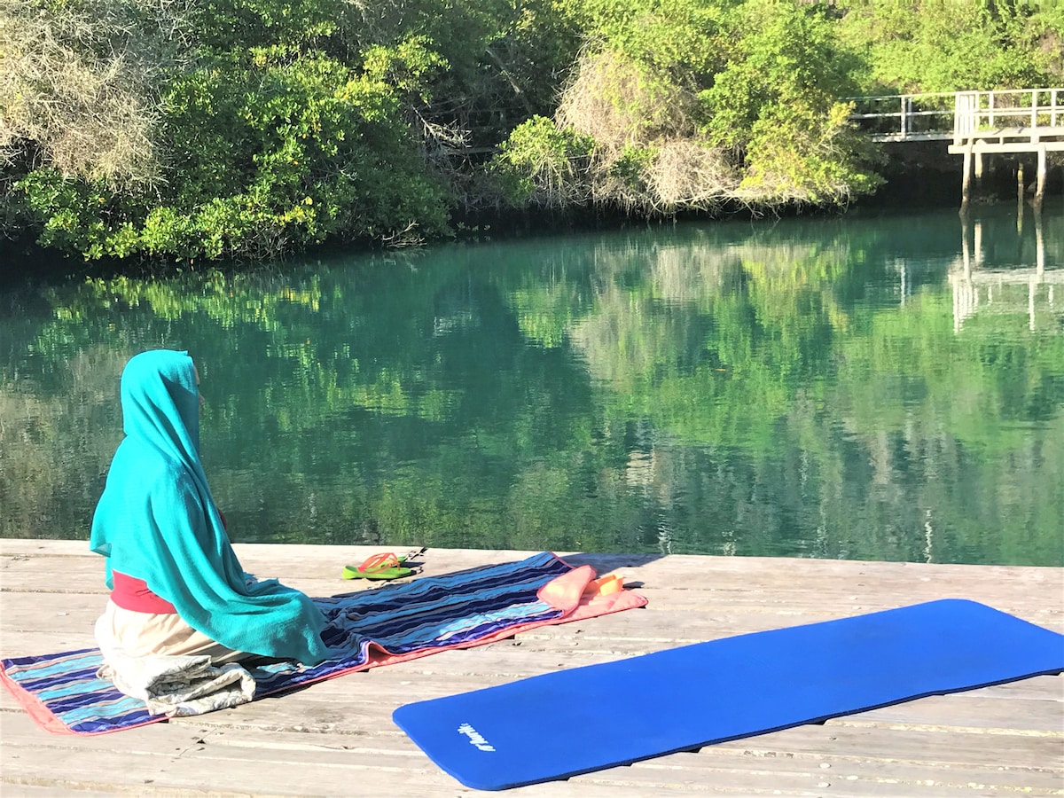 A serene individual sits peacefully on a colorful mat by the tranquil waters of Ninfas Lake. Lush greenery reflects in the clear water, enhancing the calm environment. A yoga mat is placed nearby, suggesting a space for relaxation and meditation in nature.
