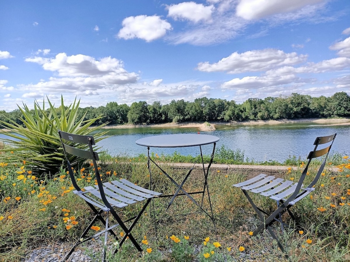 A tranquil outdoor seating area overlooks the Loire River, featuring a round table and two striped chairs. Surrounding wildflowers add a splash of color to the natural landscape, while the serene water reflects the clear blue sky dotted with clouds.