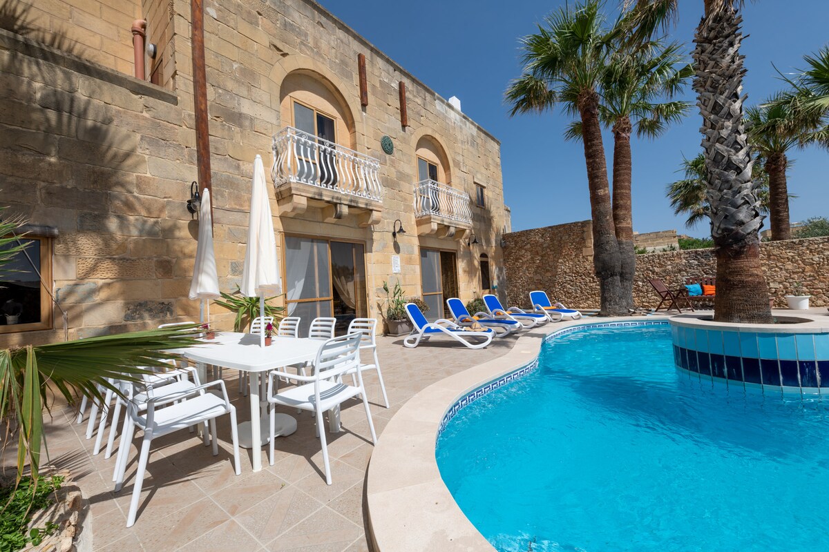 A spacious outdoor area is showcased, featuring a pool surrounded by lounge chairs under the sun. A dining set is positioned nearby, complemented by palm trees and a stone wall, creating an inviting atmosphere for relaxation and leisure.