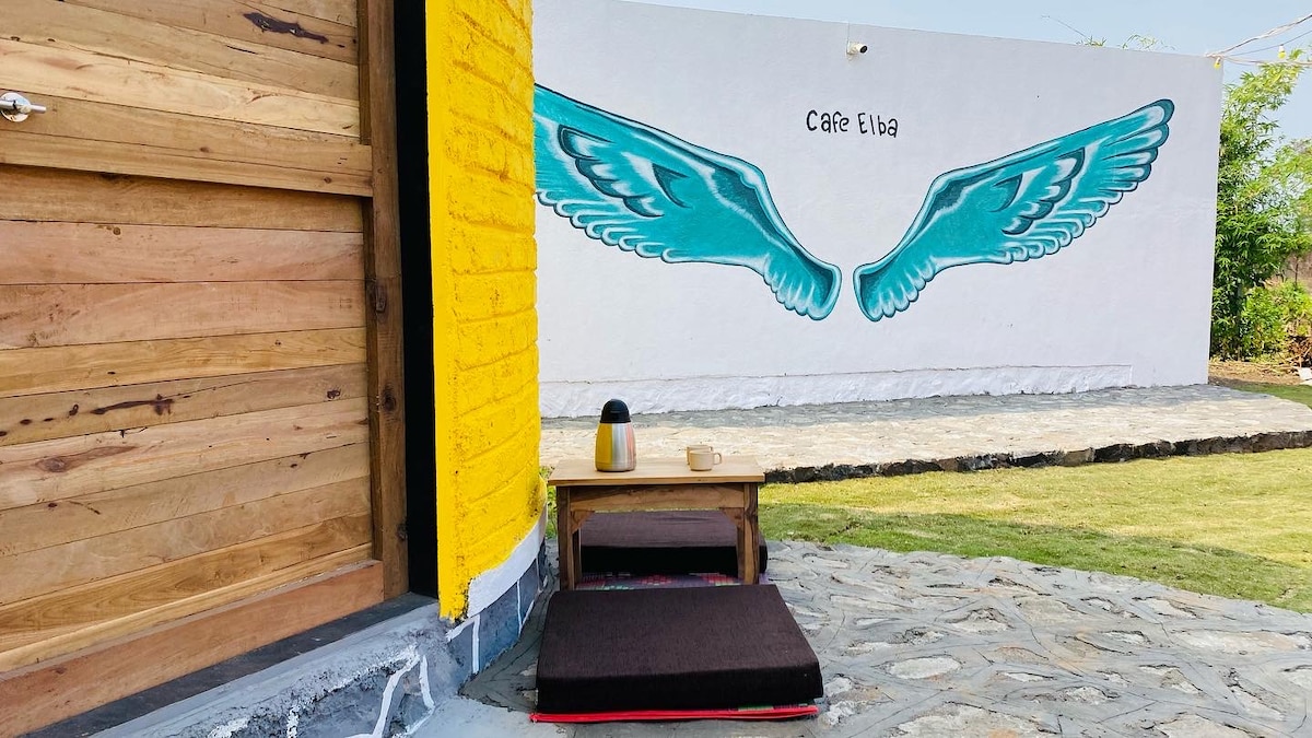 A vibrant mural of angel wings decorates an exterior wall, contrasting with the yellow paint of the building. A wooden table is positioned beside a low seating area with cushions, set on a stone pathway that leads to a grassy space.