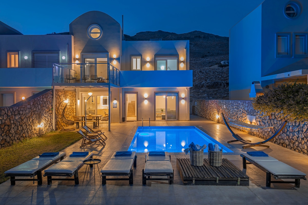 A well-lit outdoor area is highlighted by a private swimming pool, surrounded by sun loungers. A wooden deck with seating is positioned nearby. The villa's two-story facade displays large windows and a circular feature above the entrance, set against a mountain backdrop under evening light.