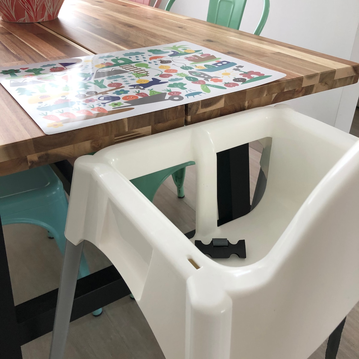 A wooden dining table with a smooth, polished surface is paired with colorful chairs. A high chair is positioned beside the table, featuring a simple white design. A playful placemat is displayed on the table, covered in various bright illustrations, adding a cheerful touch to the setting.