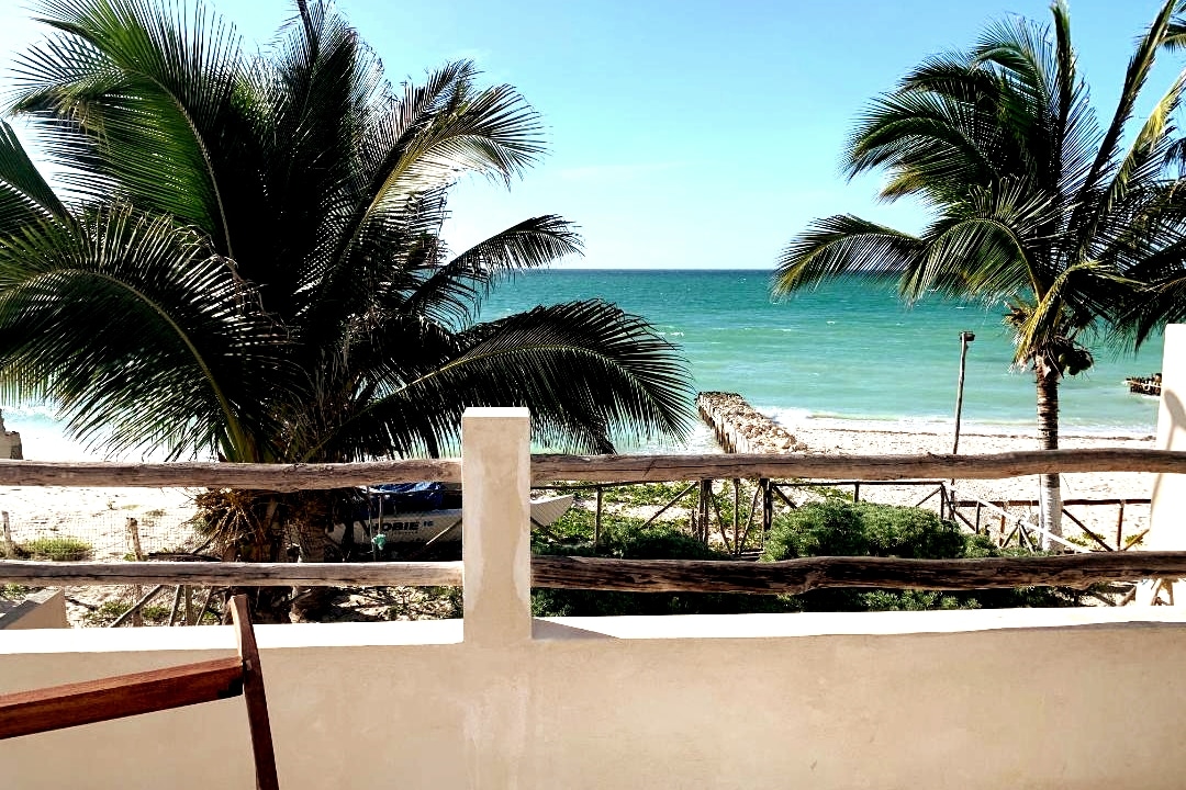 A view of the ocean is framed by palm trees from a private beachfront terrace. Sunlight reflects off the water, creating a serene atmosphere. The terrace railing is made of wood, enhancing the natural environment. Sandy beach is visible in the foreground.