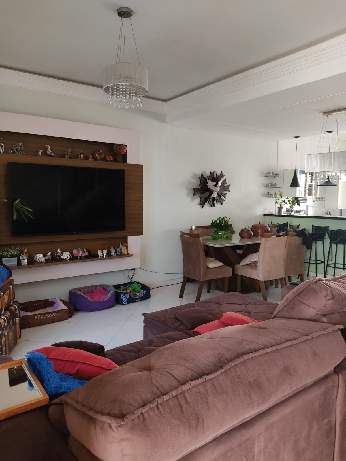 A living area features a large wall-mounted television above a wooden panel, complemented by decorative items. Nearby, a dining space is highlighted by a table surrounded by four upholstered chairs. Soft furnishings in the seating area include colorful cushions on a plush sofa.