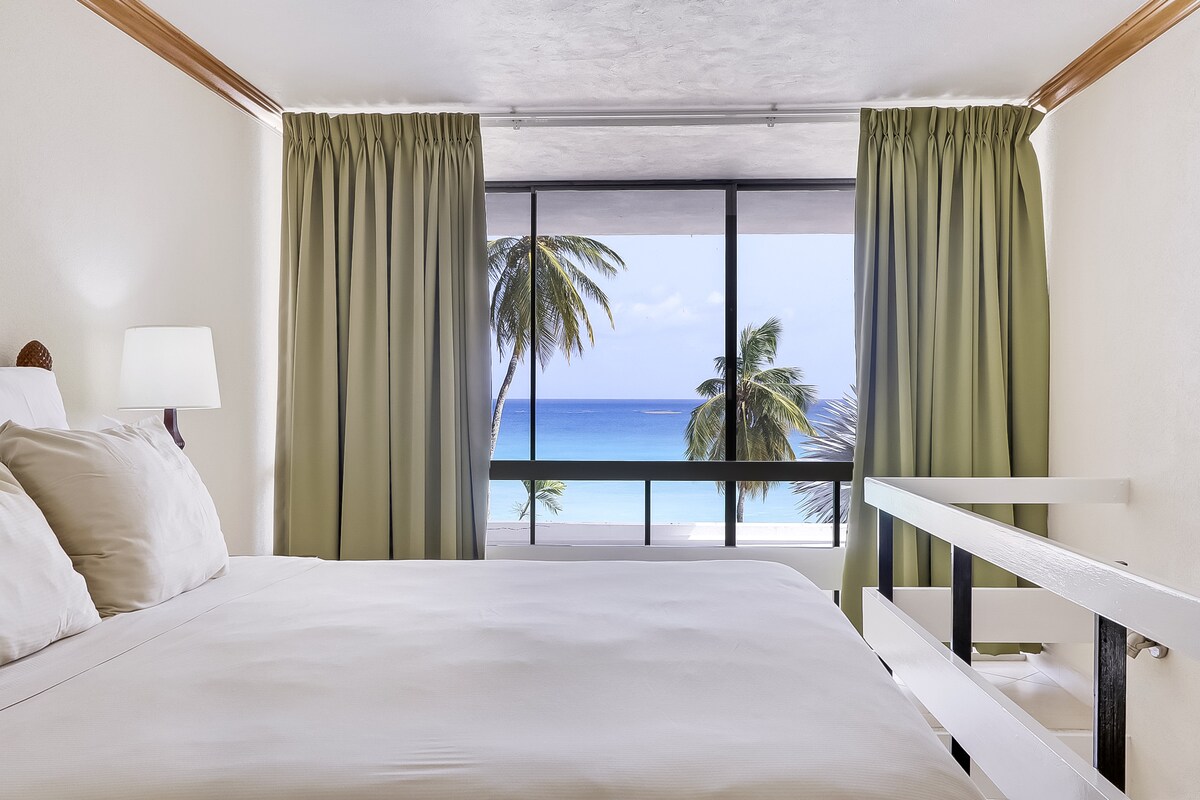 A bright sleeping area is shown with a spacious bed adorned with soft pillows. Large windows dressed with light green curtains offer unobstructed views of the ocean and palm trees, allowing natural light to fill the room.