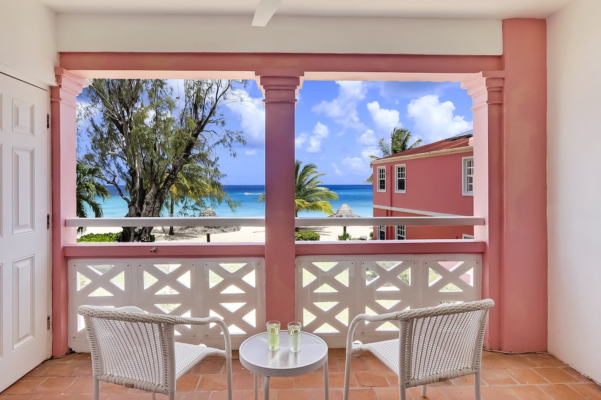 A private balcony is furnished with two white wicker chairs and a small table, offering ocean views. The scenery features a sandy beach and palm trees under a bright blue sky filled with clouds.
