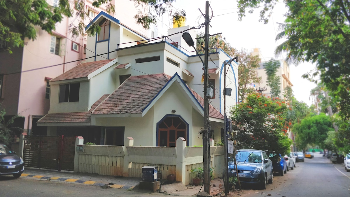 A charming duplex house is featured, showcasing a pitched roof and a mix of white and blue exterior. Lush greenery surrounds the property, and a quiet street lined with parked cars adds to the neighborhood's residential ambiance.