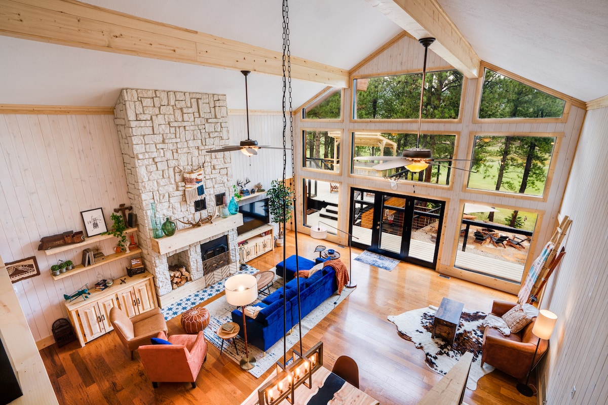 A spacious living area is highlighted by a stone fireplace and large windows that offer views of the outdoor deck and surrounding trees. Comfortable seating is arranged around the fireplace, with a dining table in the foreground and wooden accents throughout the space.