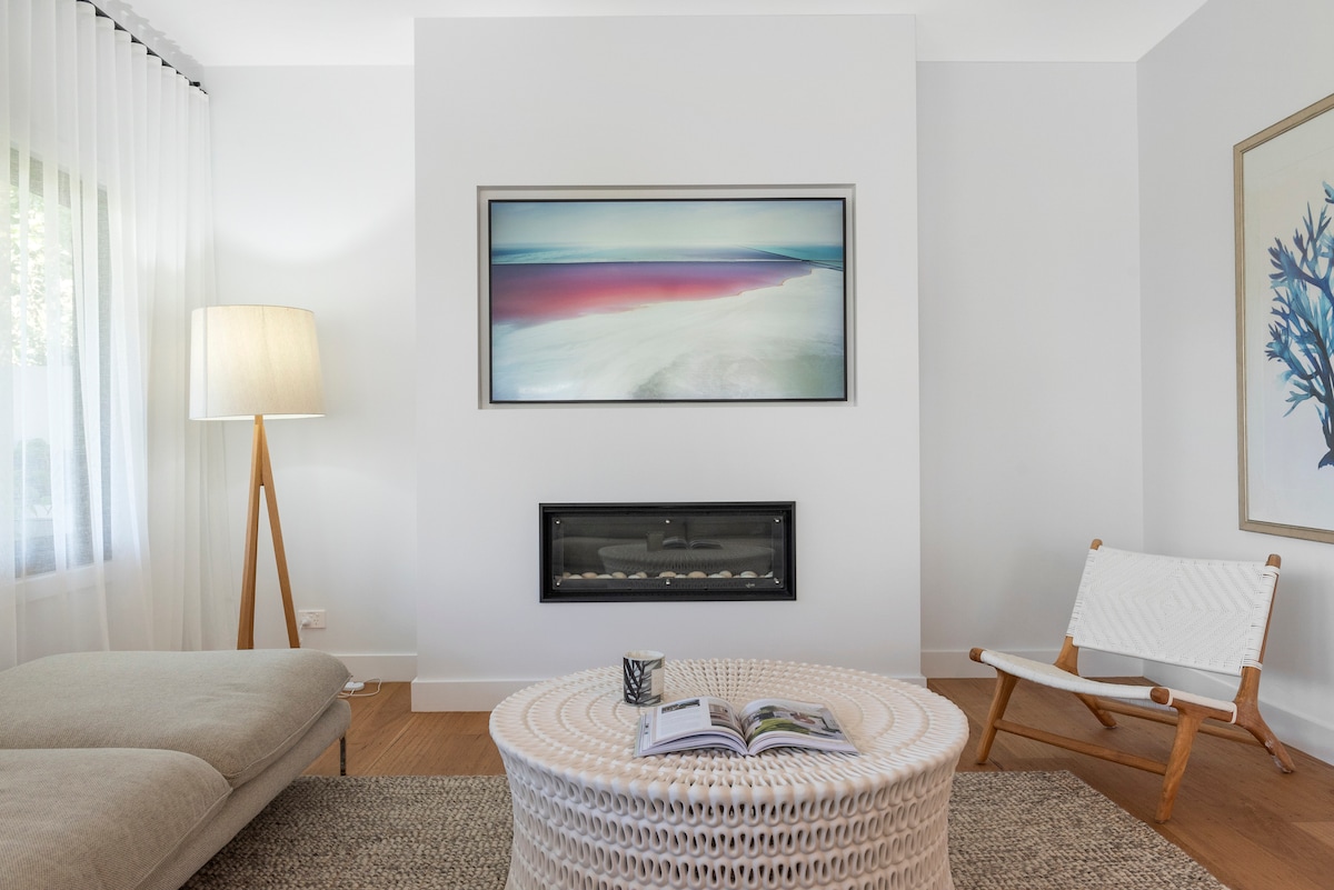 A cozy living area features a modern light grey lounge and a white woven coffee table. A framed artwork depicting abstract ocean views is displayed on the wall above the gas fireplace. Soft natural light streams through sheer white curtains, enhancing the relaxed ambiance.