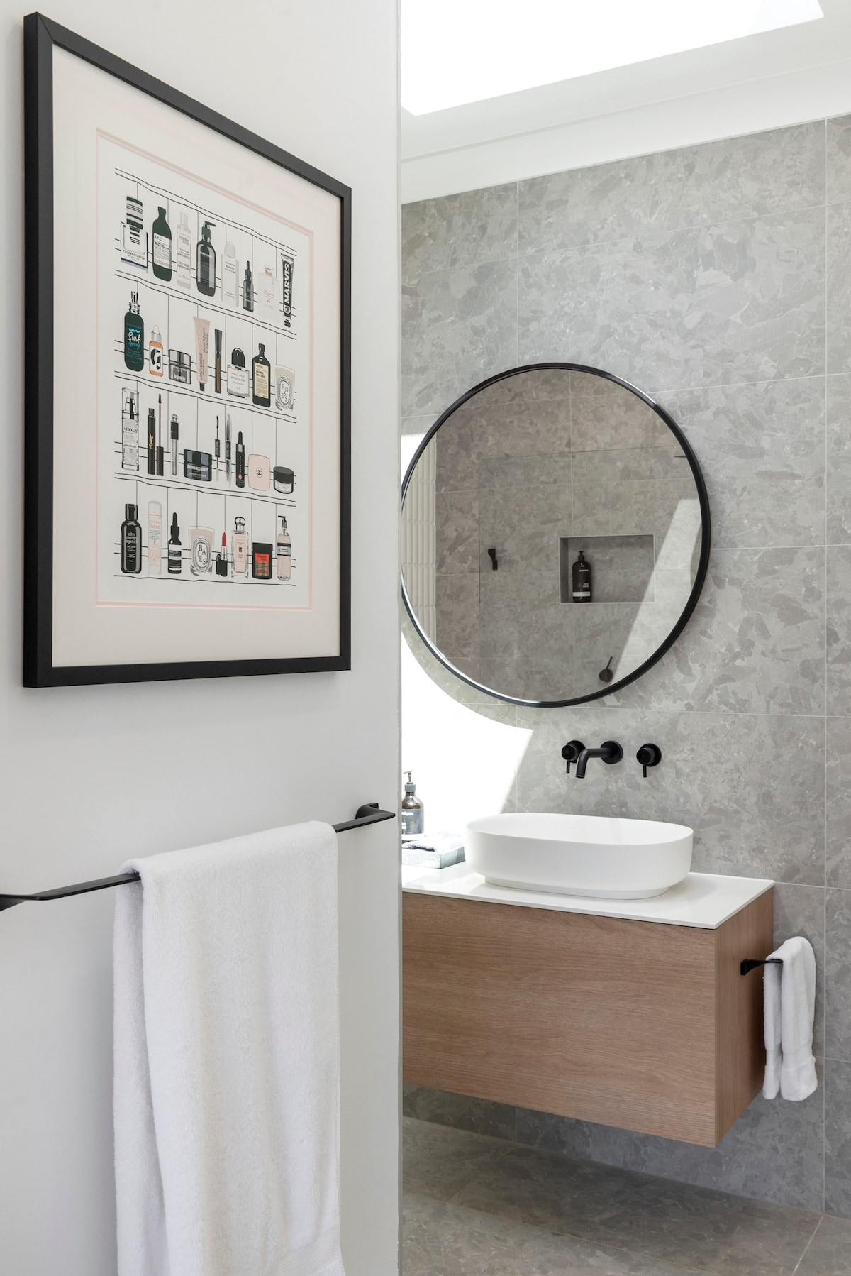 A modern bathroom features a circular mirror above a white vessel sink, complemented by a wooden vanity. Stylish art is displayed on the wall, and a white towel is neatly hung on a black towel rack. Natural light brightens the space through a skylight.