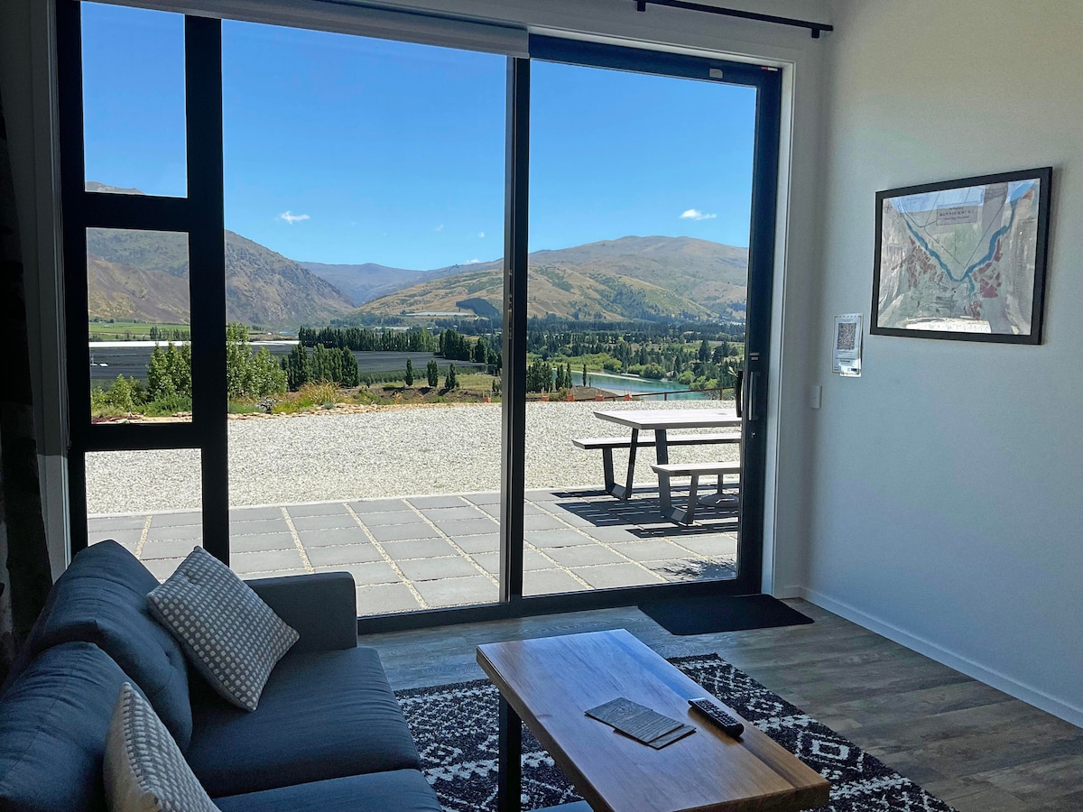 A lounge area with a comfortable gray sofa is positioned near a large glass door, providing an unobstructed view of the scenic landscape featuring hills and valleys. A wooden coffee table sits in front of the sofa, while an outdoor dining area is visible through the door.