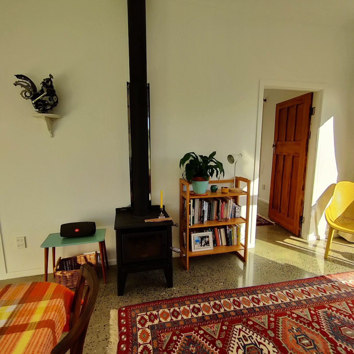 The open living area features a modern wood stove against a minimalist wall, providing warmth. A vibrant area rug adds color underfoot, while a small bookshelf holds various books and a plant is placed nearby. Light streams in through an adjacent door, enhancing the space.