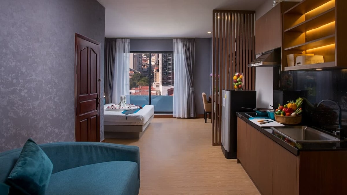 A modern studio space is presented, featuring a comfortable seating area with a blue couch. A spacious bed is positioned near a large window, allowing natural light to illuminate the room. A kitchenette with wooden cabinetry showcases fresh fruit on the counter.