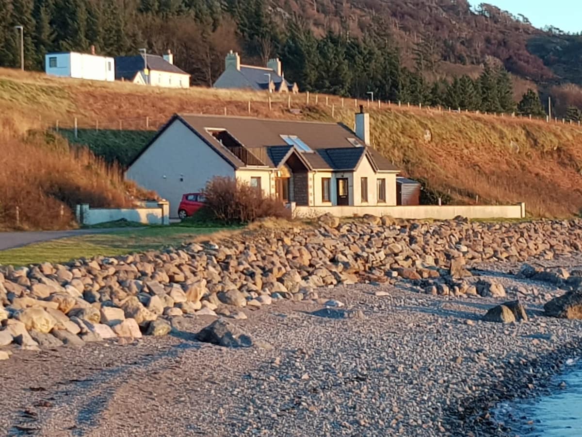 A modern house is positioned along a pebbled shoreline, with grassy slopes rising behind it. Large windows reflect the natural light, creating a harmonious connection with the landscape. The front yard features a low stone wall, enhancing the serene coastal setting.