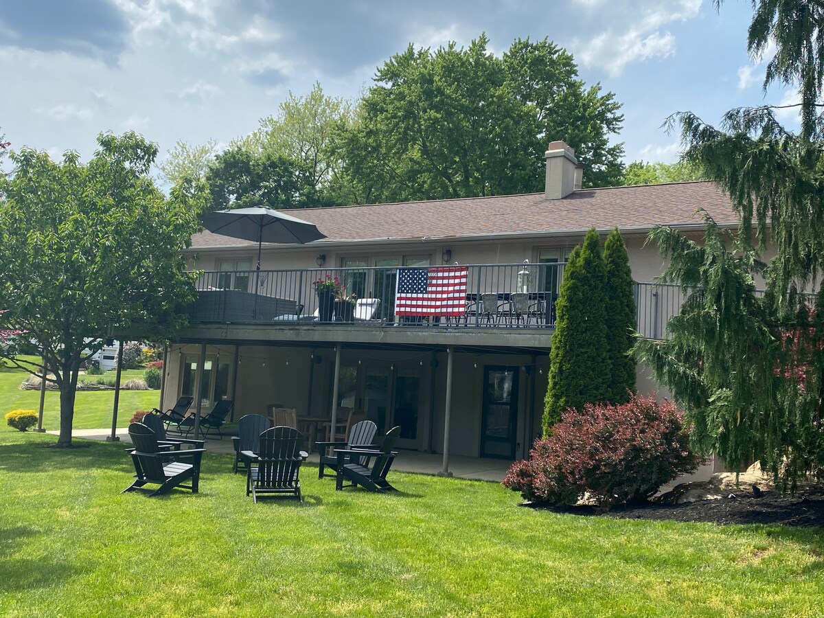The exterior of a two-level home is visible, featuring a large balcony adorned with an American flag. The well-manicured lawn includes several Adirondack chairs arranged around a firepit, surrounded by lush trees and shrubs, creating a relaxing outdoor environment.