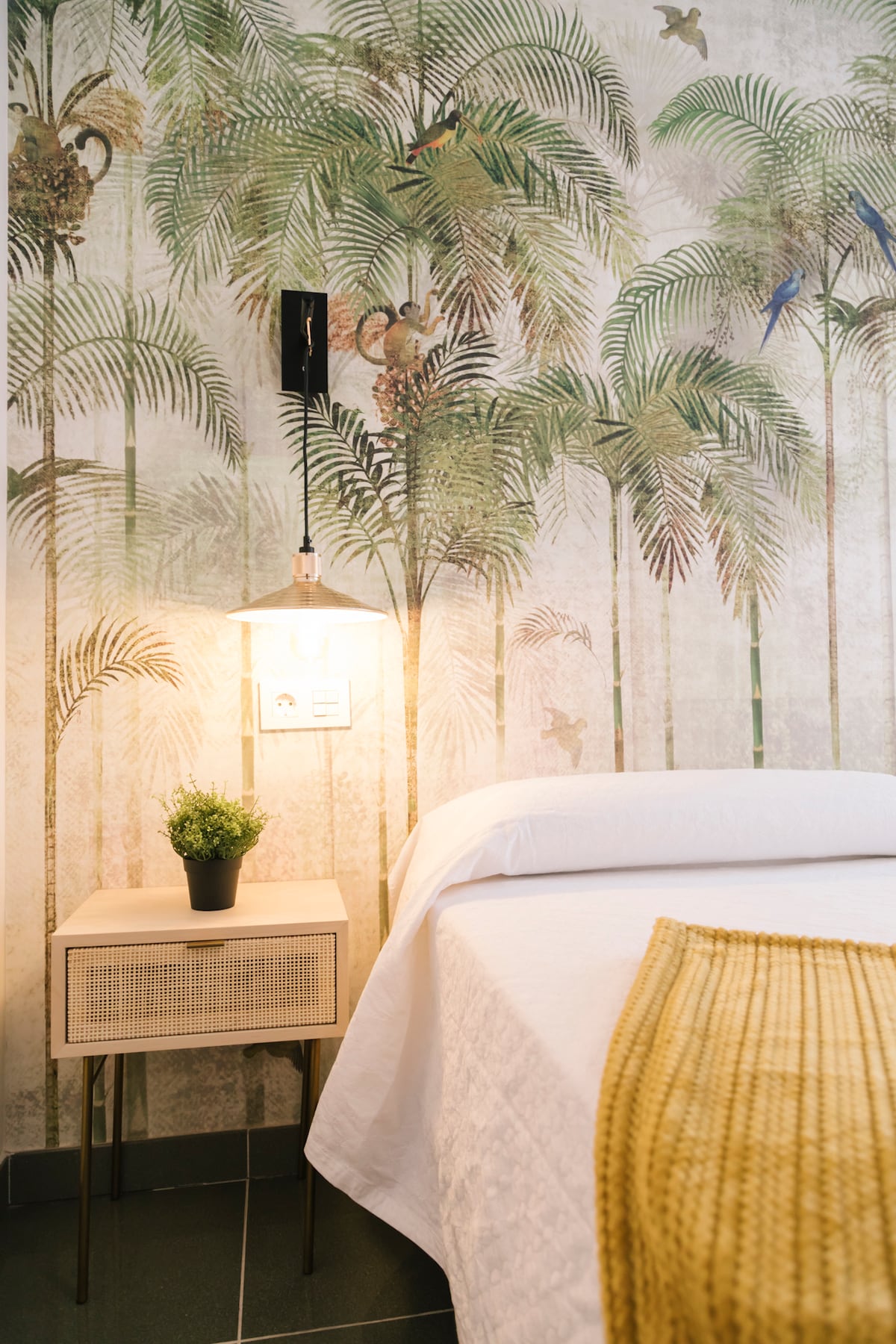 The sleeping area features a light-colored bed against a vibrant tropical wallpaper adorned with palm trees and birds. A small nightstand holds a potted plant, and a pendant light provides soft illumination. A textured yellow throw lies across the bed, enhancing the space's warmth.
