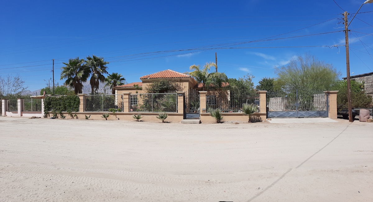 The exterior of the house is showcased, featuring a warm-toned stucco facade with a clay tile roof. Lush palm trees and desert vegetation frame the property, which is enclosed by a decorative fence. A clear blue sky adds to the inviting outdoor ambiance.
