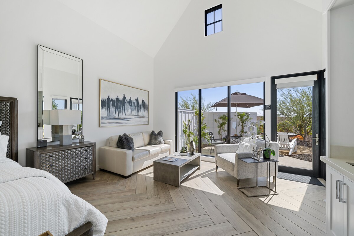 A spacious living area is highlighted by high ceilings and large windows that invite natural light. A comfortable sofa faces a coffee table, while a side chair complements the seating arrangement. Outside, a patio with an umbrella and landscaped greenery is visible.