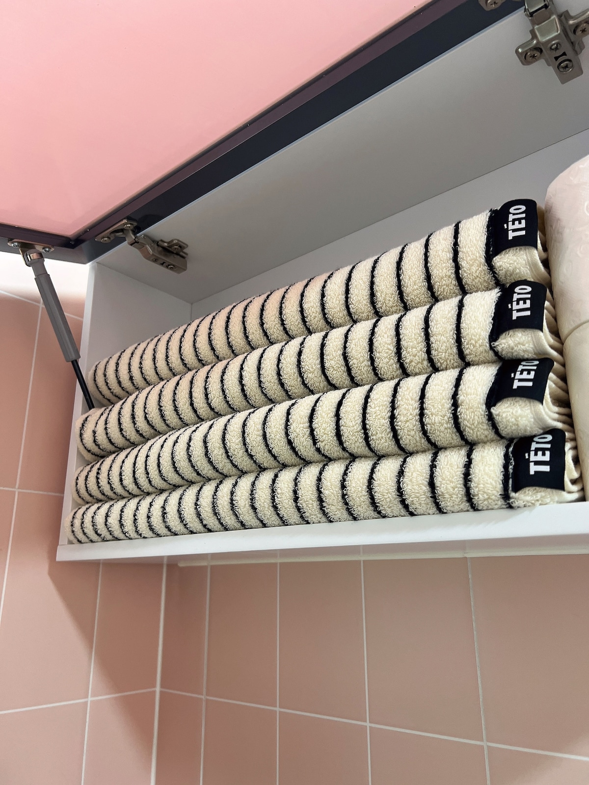 A neatly organized shelf is filled with several rolled towels, displaying a striped pattern. The towels are stacked uniformly, showcasing their light color against the soft pink tiles of the bathroom.