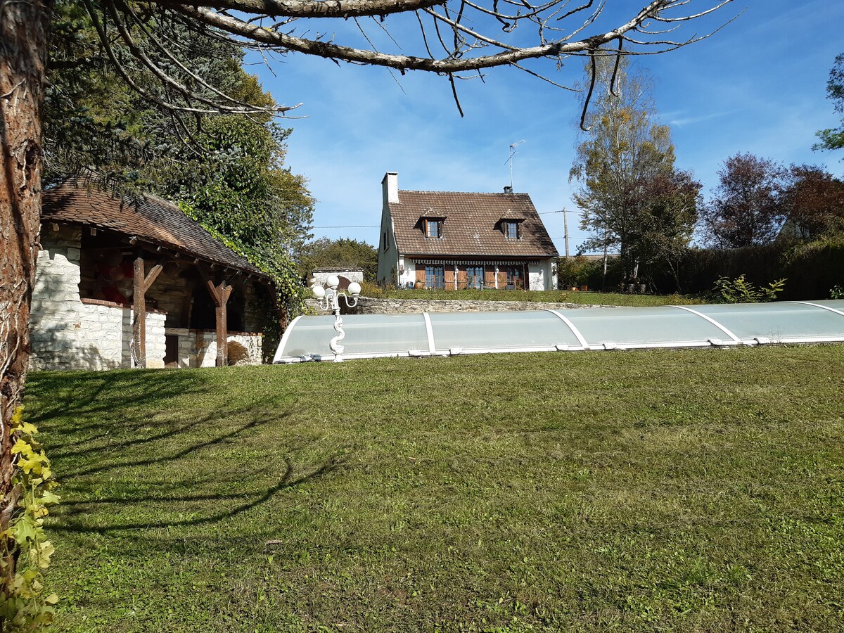 A spacious garden area is depicted, featuring a well-maintained lawn bordered by trees. In the background, a rustic house with a sloped roof and multiple windows is visible. A covered swimming pool is positioned to one side, alongside an outdoor structure that may serve as a relaxation or cooking area.