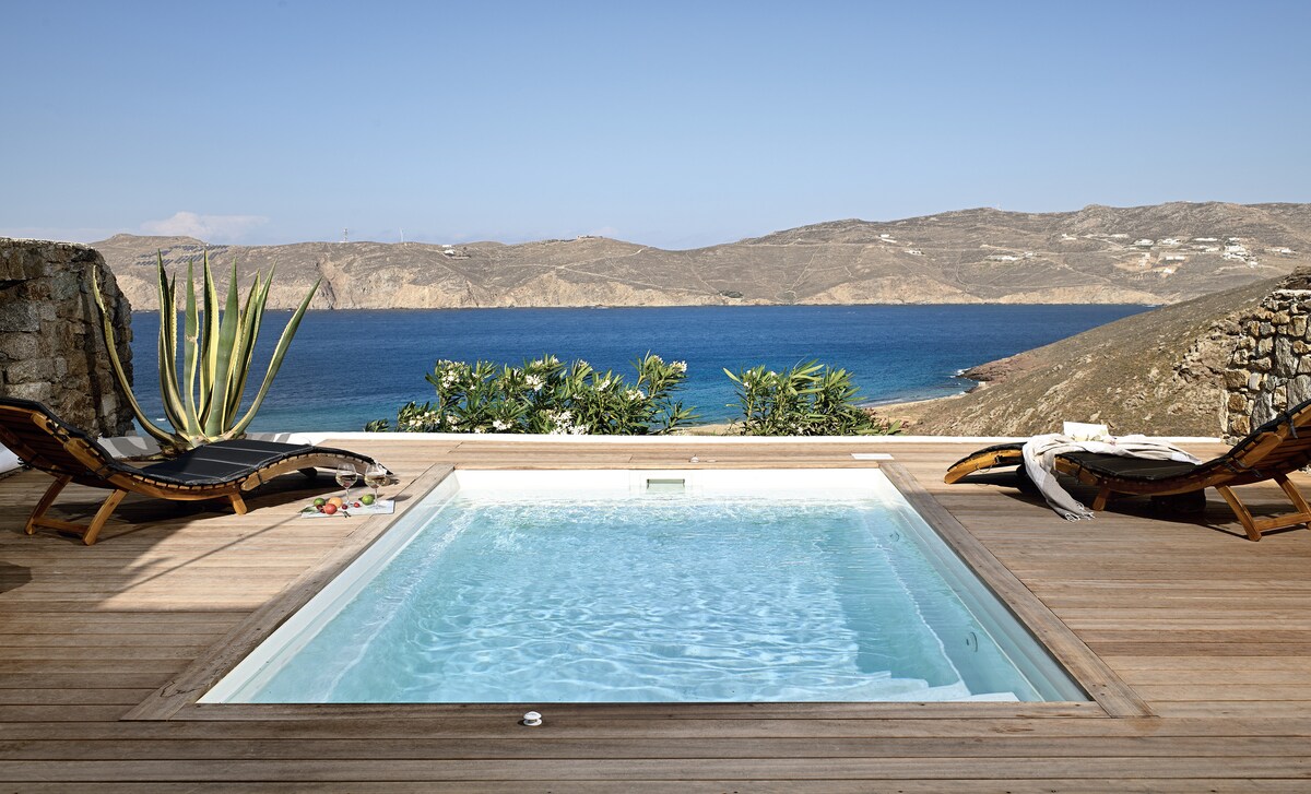 A private pool is featured with a clear blue surface that reflects the sky. Two sun loungers, positioned beside the pool, offer views of the distant hills and the sparkling ocean. Lush greenery surrounds the area, enhancing the natural setting.