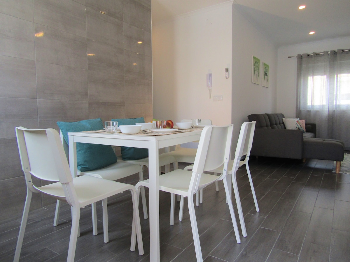 A modern dining area features a white table surrounded by six white chairs. Natural light enters through a nearby window, complementing the light grey tiled floor and warm wall tones. A cozy seating area with a dark grey sofa is visible in the background.