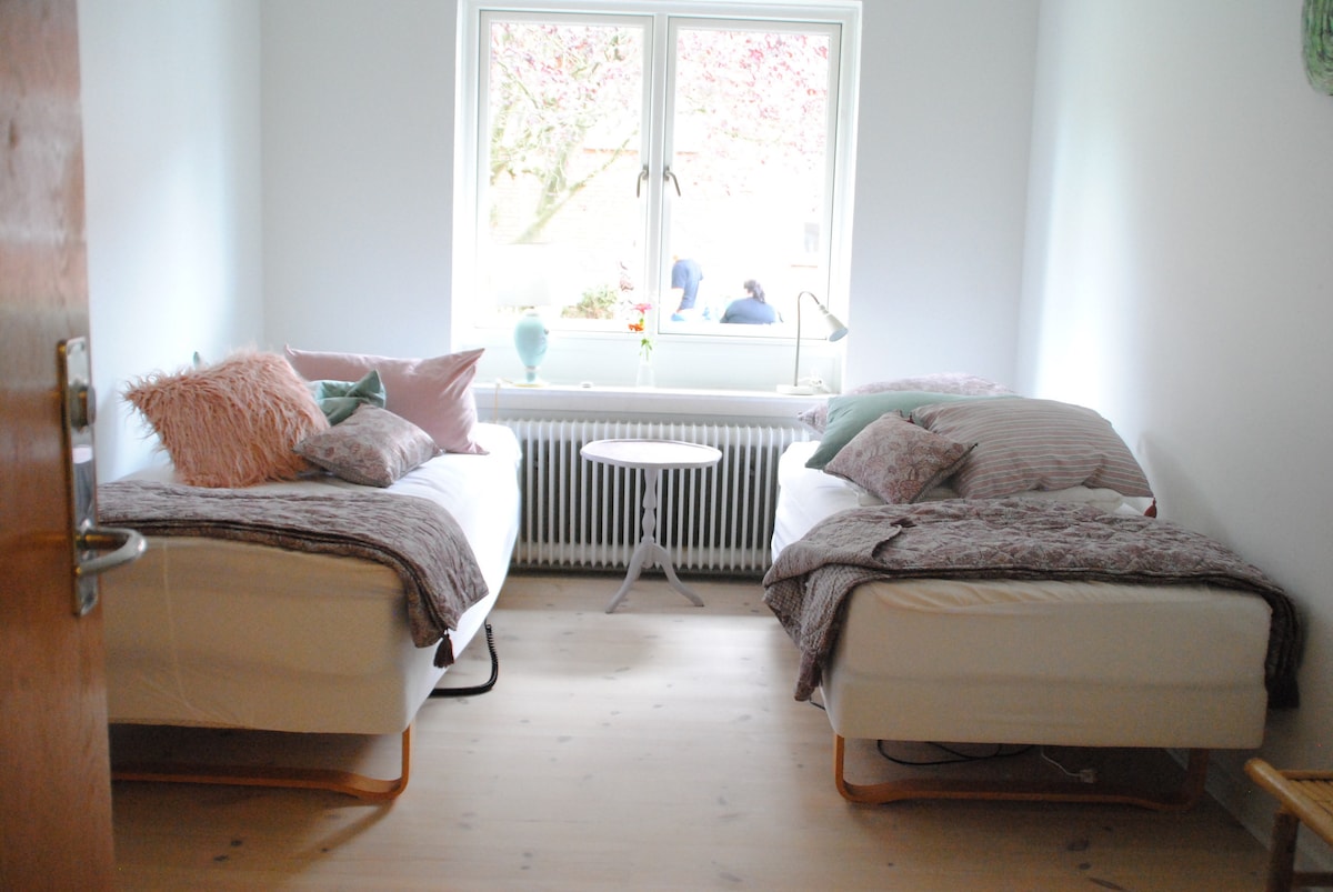 A bright bedroom features two twin beds dressed in soft linens, with decorative pillows adding subtle color. A small round table is positioned between the beds, and natural light enters through a large window overlooking a garden.