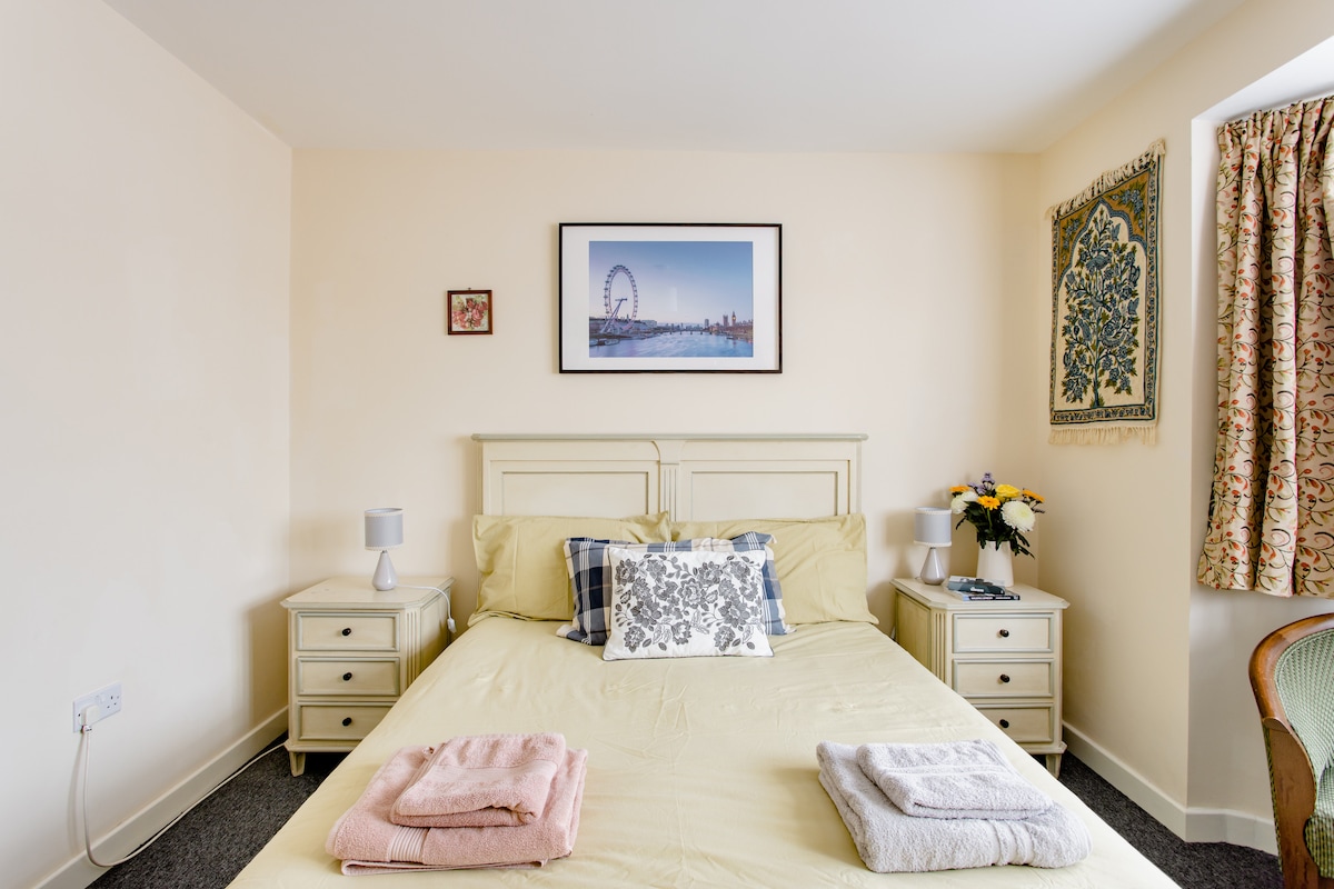 A spacious room is furnished with a king size bed, complemented by bedside tables and lamps on either side. A framed photograph hangs above the bed, depicting a cityscape, while decorative curtains and wall art add a touch of color to the neutral decor.