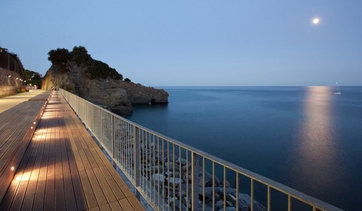 A wooden boardwalk stretches along the coastline, providing a serene view of the calm sea under a moonlit sky. The soft glow of ground lights illuminates the pathway, with rocky cliffs visible on the left, offering a tranquil atmosphere for evening strolls.