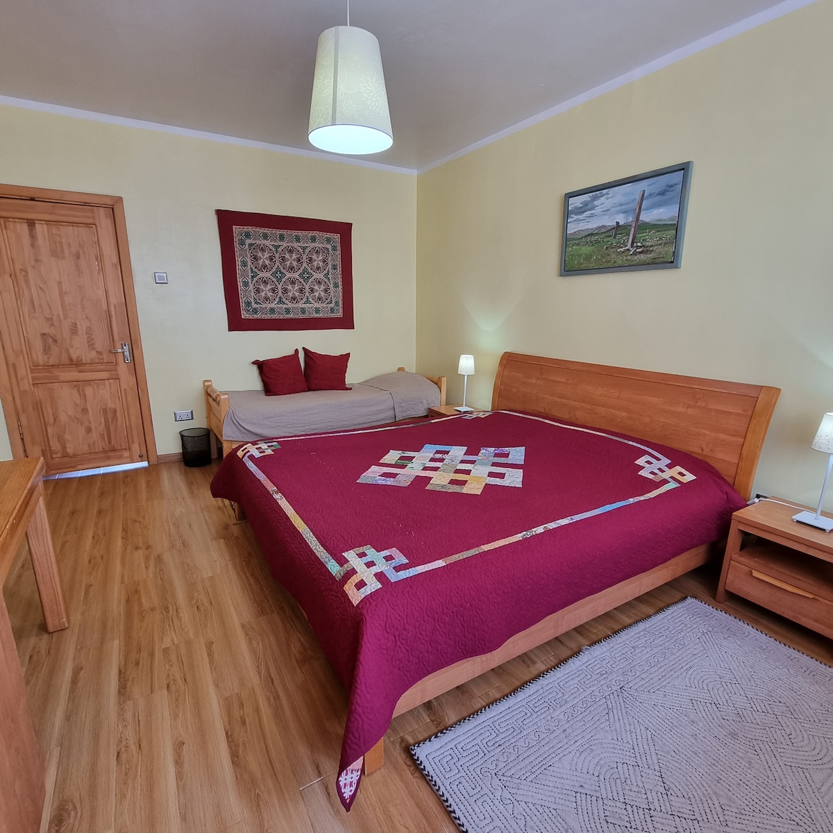 A spacious bedroom features wooden furniture, including a large bed covered in a deep red quilt with a geometric pattern. A smaller bed is positioned beside it. Soft lighting is provided by bedside lamps, and vibrant artwork adorns the walls.