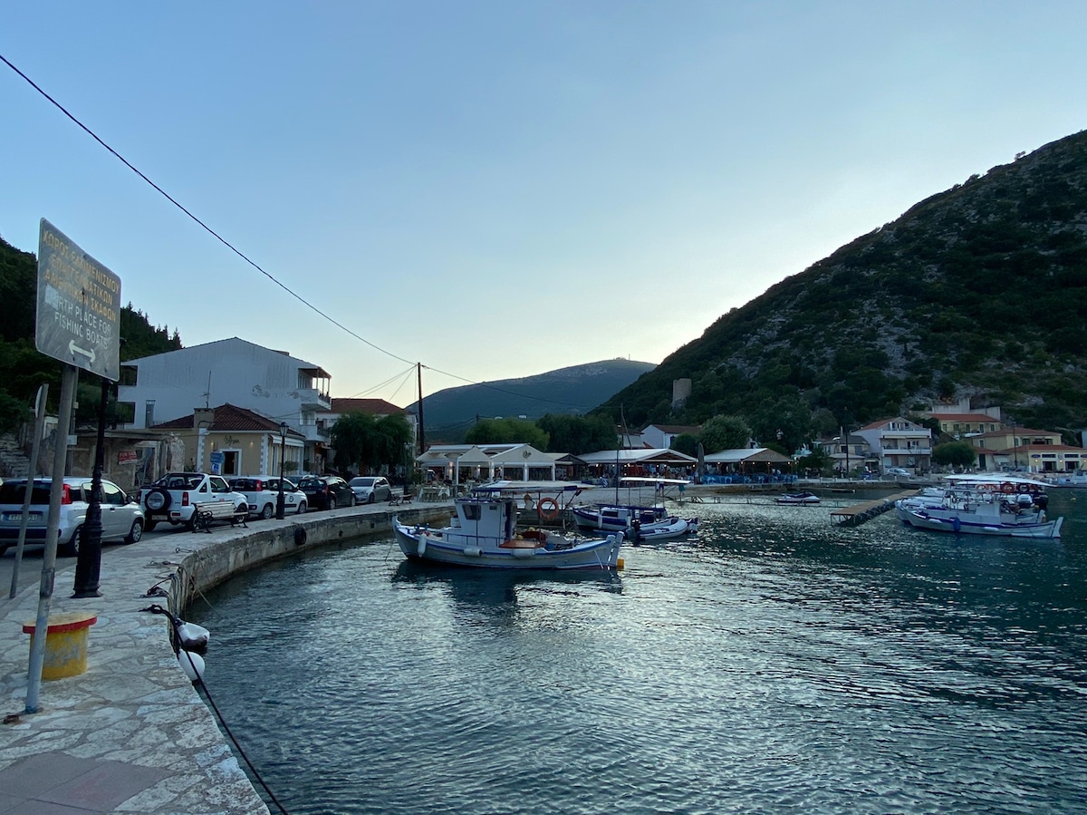 A tranquil seaside setting features a harbor lined with small fishing boats. Nearby, a road is visible with parked cars and buildings that include dining establishments. The surrounding hills provide a scenic backdrop, complementing the calm waters and evening light.