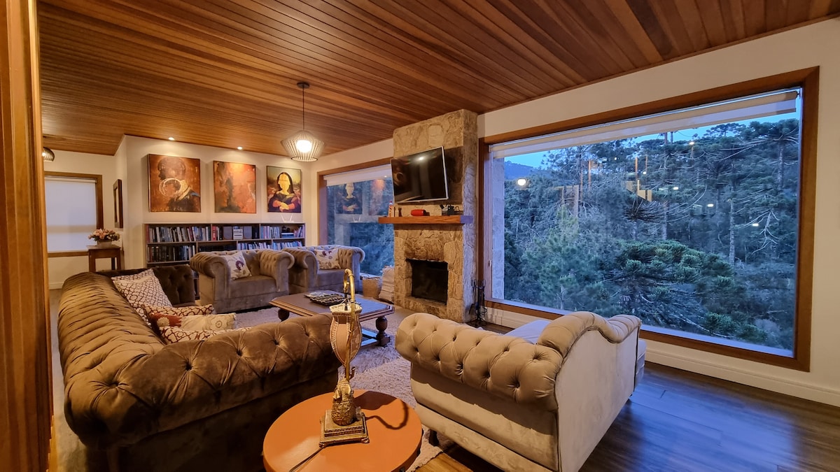 A spacious living room features a cozy seating arrangement with tufted sofas arranged around a central coffee table. Large windows offer views of the surrounding greenery, while a television and a stone fireplace add to the inviting ambiance.