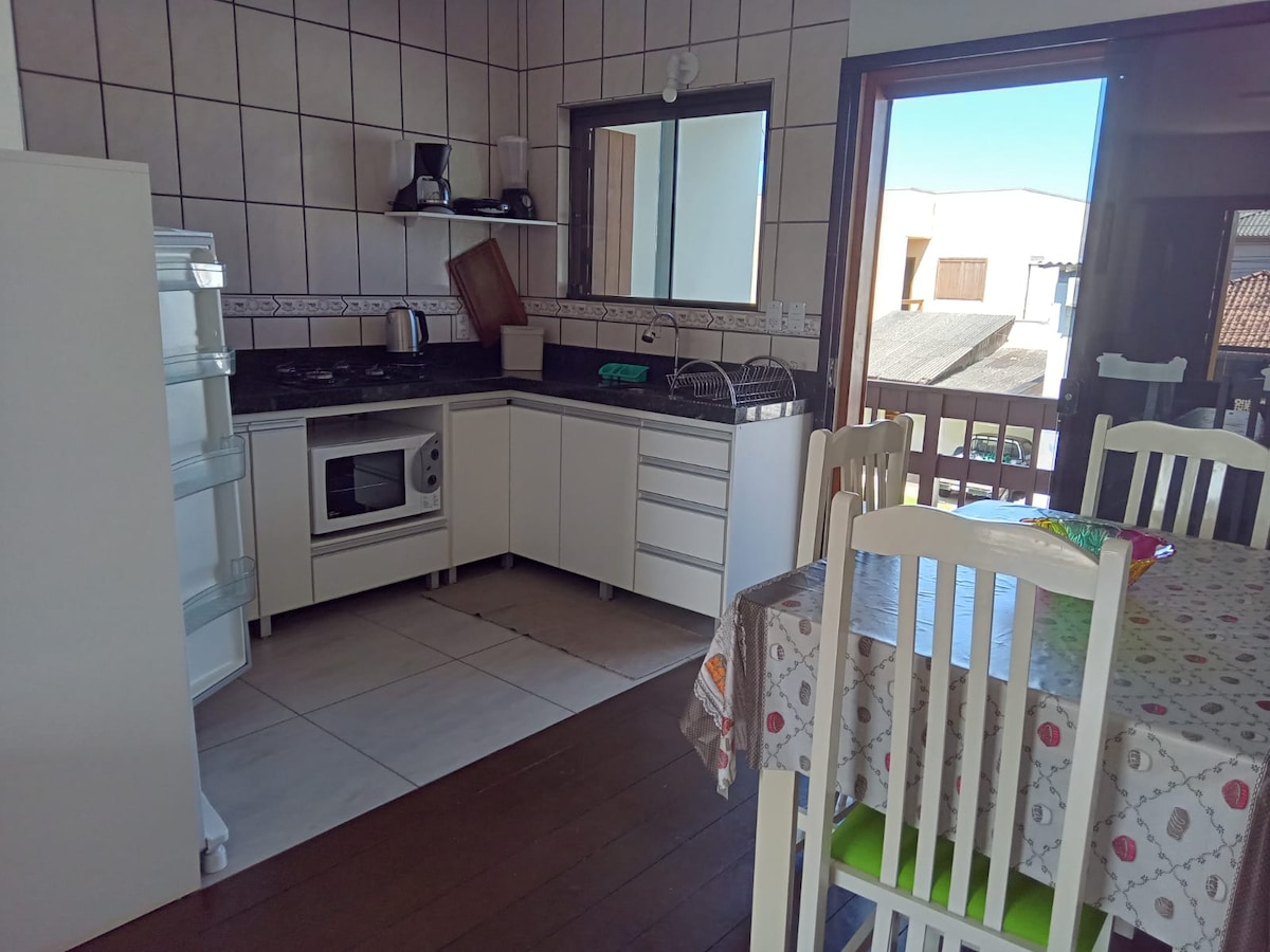 The kitchen area displays modern cabinetry and appliances, including a refrigerator, microwave, and gas stove. A large window provides natural light, complementing the dining table set for six. The floor features a mix of tiles and wood, creating a functional and welcoming space.