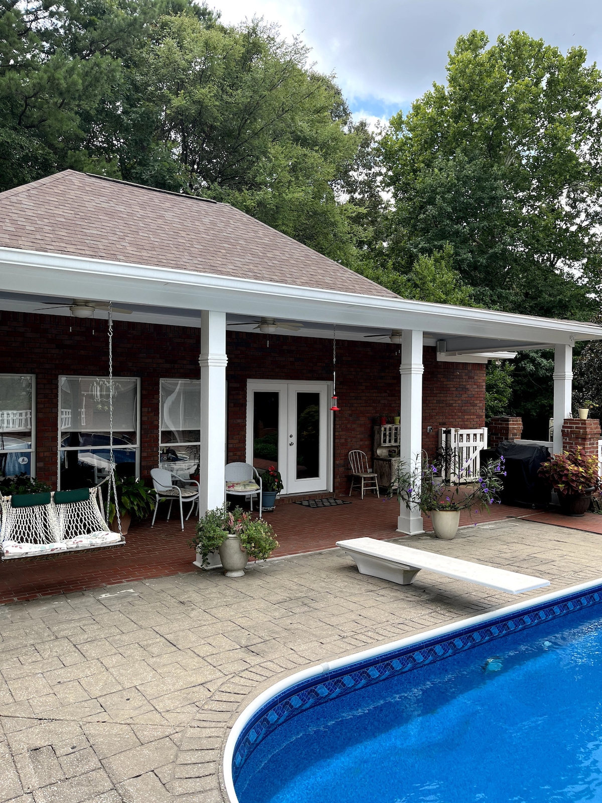 An inviting poolside area is shown with a sparkling blue pool in the foreground. The spacious covered patio features white pillars and is furnished with a swing and several seating options. Lush greenery surrounds the area, enhancing a relaxed outdoor atmosphere.