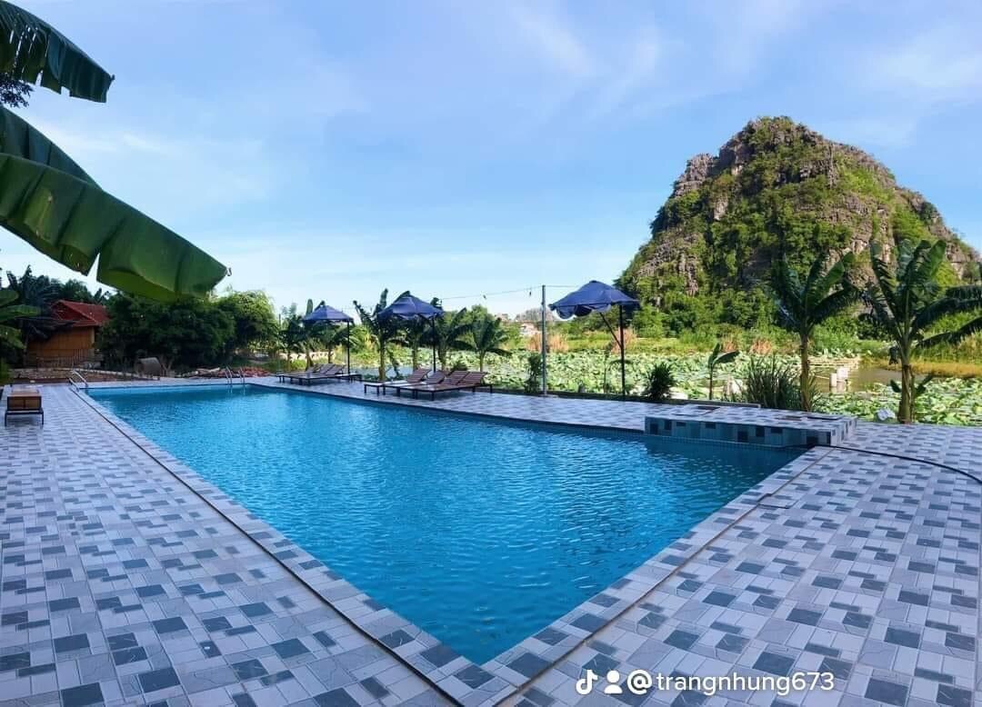 An inviting outdoor pool is surrounded by lush greenery and palm trees, with blue skies overhead. Comfortable lounge chairs and umbrellas are positioned around the pool area, offering space for relaxation. A scenic mountain view is visible in the background, enhancing the tranquil setting.