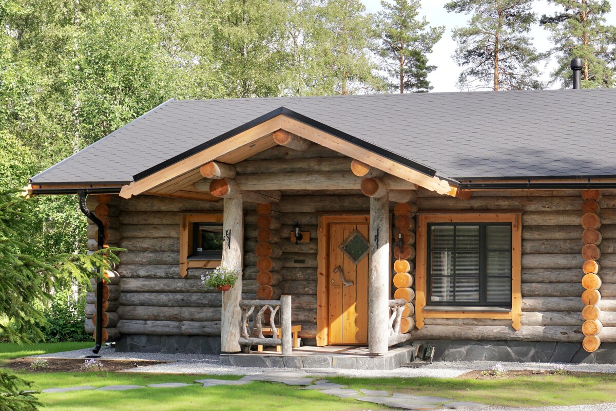The exterior of the cabin is constructed from hand-carved kelo logs, featuring a sloped roof and wooden accents. A covered porch with seating invites relaxation. A well-kept lawn surrounds the house, complemented by trees in the background that enhance the natural setting.
