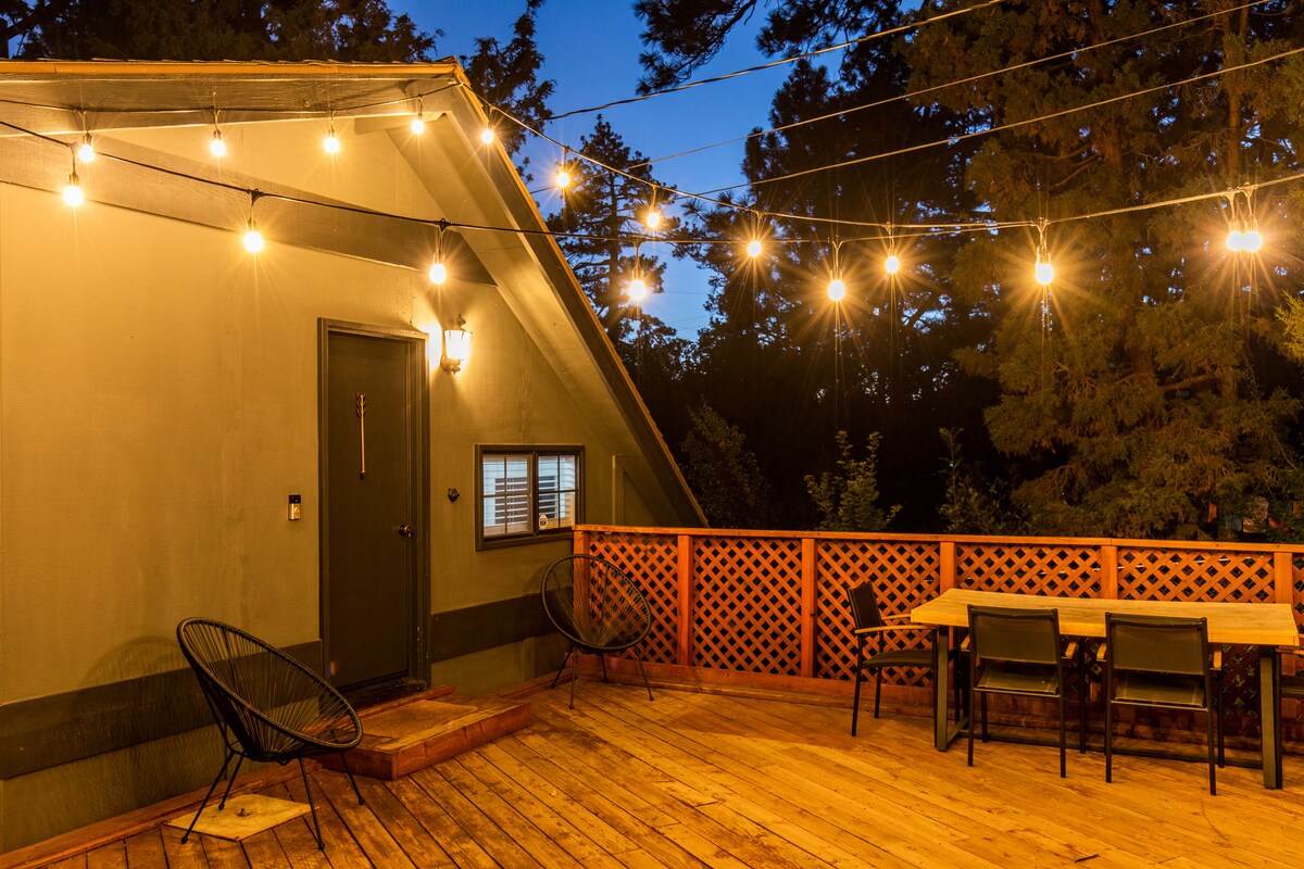 A spacious deck is illuminated by string lights, creating a gentle glow in the evening. The deck features a wooden table with seating for four, as well as two stylish lounge chairs. A single door leads into the guesthouse, surrounded by a natural setting.
