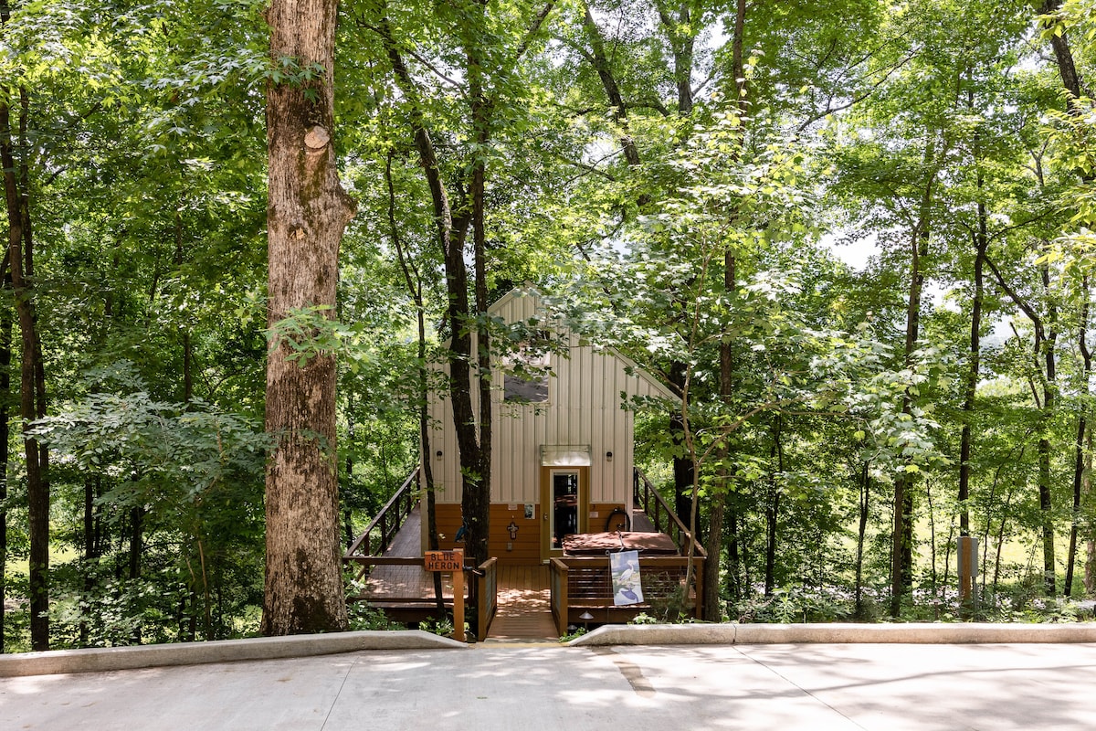 The cabin is nestled among lush green trees, with a welcoming entrance leading to a deck. A pathway extends from the driveway to the cabin, offering a serene, natural atmosphere. Light filters through the surrounding foliage, enhancing the secluded setting.